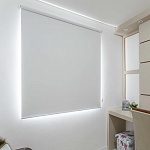 Persiana Rolô Blackout Branca em uma parede branca com alguns móveis típicos de quarto dispostos nos cantos da imagem